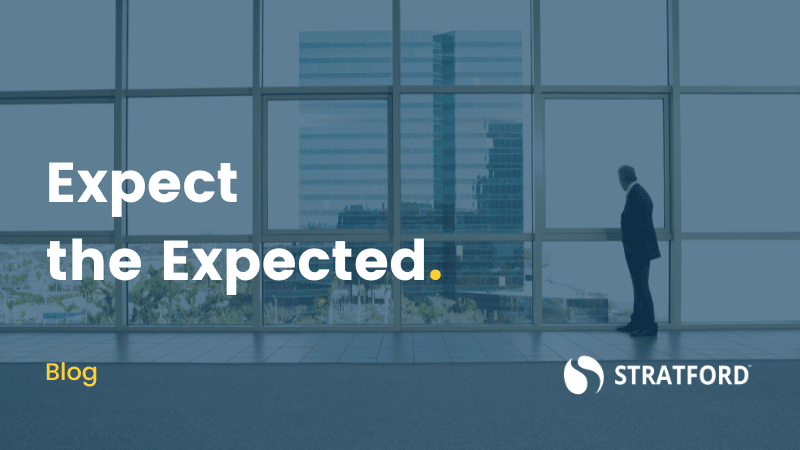 Wide shot of businessman in suit standing in front of floor-to-ceiling windows surveying skyline. Text overlay "Expect the Expected."